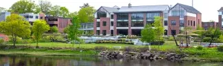 A large brick building with large windows sits on the edge of a river bank surrounded by green trees and grass